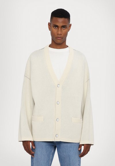 Cardigan créme avec col et poignets côtelés, dotée d'une fermeture à boutons et de deux poches avant; motif en tricot texturé, coupe décontractée.