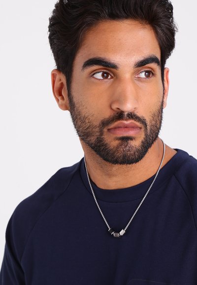 Zilveren ketting met zwarte en metallic accenten, gedragen door een persoon in een donkerblauw shirt met een ronde hals en korte mouwen.