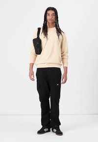 Beige sweatshirt, black cargo pants with pockets, black crossbody bag, and black sneakers with white stripes, featuring long braided hair.