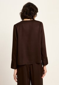 Frau mit schulterlangem, dunklem Haar, die ein lockeres, langärmliges, dunkelbraunes Oberteil und passende Hosen trägt, von hinten gezeigt.