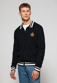 Chaqueta negra de punto con cremallera, cuello y puños de rayas blancas acanaladas, con un insignia bordada redonda en el pecho. Patrón texturizado en toda la prenda.