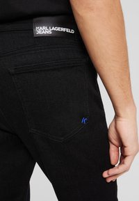 Fekete farmernadrág, bőr logó címkével, rajta a "KARL LAGERFELD JEANS" felirattal. Különleges kék díszvarrás a hátsó zseben.