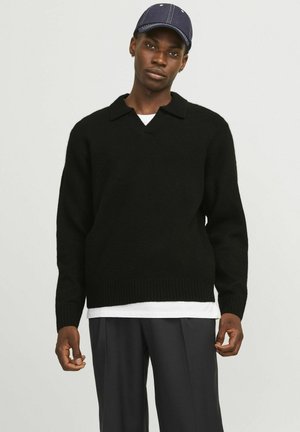 Suéter negro de punto con escote en V y cuello corto, mangas largas y dobladillo acanalado. Combinado con una camisa blanca por debajo y pantalones grises.