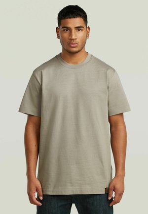 Jonge man met kort donker haar, gekleed in een effen beige oversized T-shirt met korte mouwen en donkere jeans, staand tegen een neutrale achtergrond.