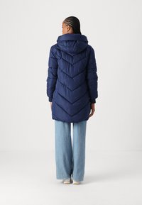 Manteau en duvet bleu marine avec capuche, présentant un motif en chevrons et des poignets côtelés, associé à un jean en denim évasé et des chaussures blanches.