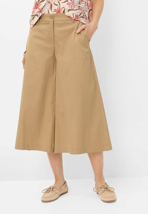 Vrouw draagt beige culottebroek met wijde pijpen, gecombineerd met beige veterschoenen en een top met bloemenpatroon, staande met één hand in de zak.