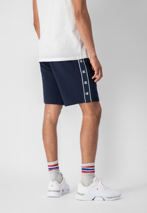 Pantaloni corti blu navy con profili bianchi e dettagli del logo. Abbinati a scarpe da tennis bianche e calzini grigi con strisce rosse e blu. Maglietta bianca.