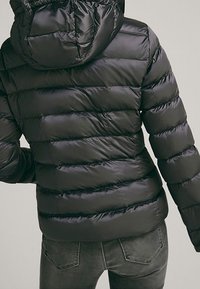 Giacca a vento nera con cappuccio, caratterizzata da sezioni trapuntate orizzontali e una texture lucida e liscia. Il design è elegante e aderente.