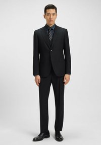 Completo nero con rever a punta, chiusura con un bottone e vestibilità slim. Abbinato a una camicia blu a fantasia e scarpe eleganti nere.