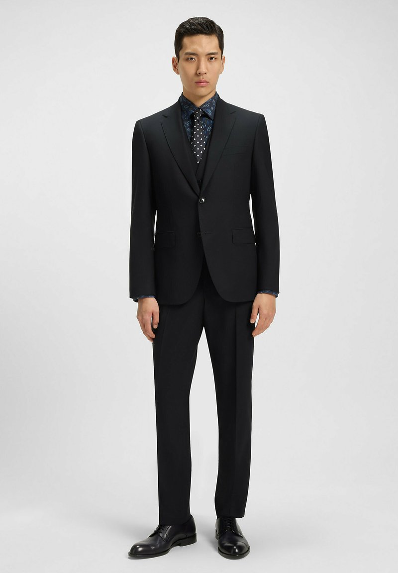 Completo nero con rever a punta, chiusura con un bottone e vestibilità slim. Abbinato a una camicia blu a fantasia e scarpe eleganti nere.