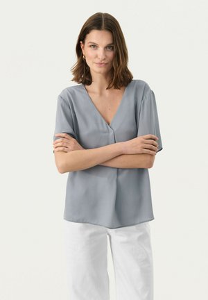 Blusa grigia chiaro con scollo a V, maniche corte e una texture liscia; abbinata a pantaloni bianchi con una vestibilità leggermente rilassata.