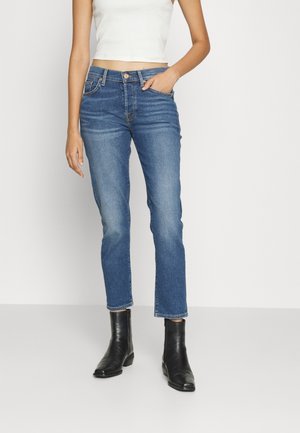 Kobieta ubrana w niebieskie jeansy o prostych nogawkach, czarne botki za kostkę i biały krótki top bez rękawów, stojąca z jedną ręką w kieszeni.