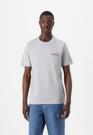 Pelēka apmaka ar apaļu kakla izgriezumu, izgatavota no kokvilnas, ar nelielu sarkanu un zilu "HILFIGER" logo uz krūšu kabatas. Īsas piedurknes, ikdienišķs piegriezums.