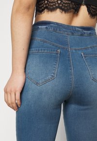 Vaqueros azules de mezclilla con una cinturilla alta, que cuentan con dos bolsillos traseros con un diseño puntiagudo, costuras oscuras y una textura suave.