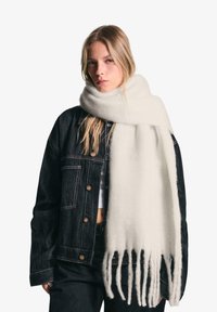 Hvid uld tørklæde med frynser, draperet omkring halsen, parret med en sort denimjakke med metalknapper og kontrastsyninger.