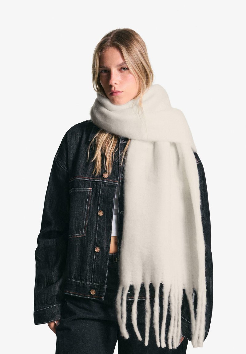 Écharpe en laine blanche avec des franges, enroulée autour du cou, associée à une veste en denim noir dotée de boutons en métal et de coutures contrastantes.