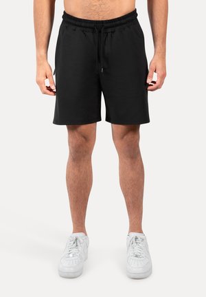 Mand iført sorte shorts med snøre og hvide sportssko, stående med armene afslappet ned langs siderne mod en ensfarvet baggrund.