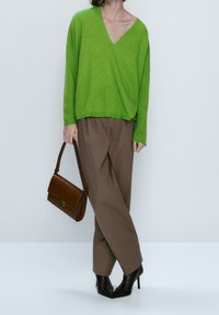 Personne portant un pull vert vif à col en V, un pantalon marron ample et des bottes à talons noires, tenant un sac à main en cuir marron à leur côté.