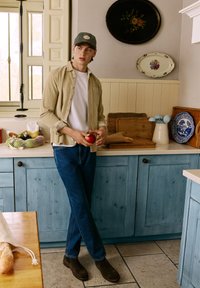 Camisa button-up de color beige claro sobre una camiseta blanca, jeans azules, botas marrones; sosteniendo una manzana roja en una cocina con armarios de madera.