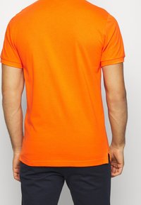 Pomarańczowa bawełniana koszulka polo z krótkim rękawem, o luźnym fasonie i bocznych rozcięciach, pokazana z tyłu.