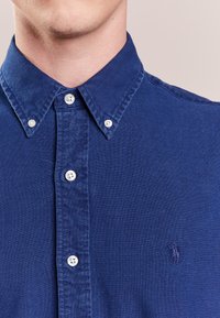 Blå button-up skjorta gjord av texturerad tyg, med spridd krage, vita knappar, och en broderad logotyp på bröstet.