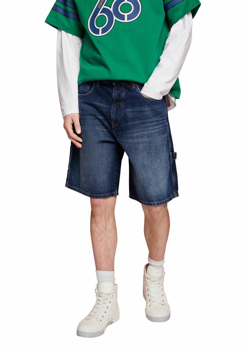 Short en jean bleu foncé, présentant une coupe décontractée, des poches latérales et un ourlet légèrement effiloché. Associé à un t-shirt graphique vert et des baskets blanches.