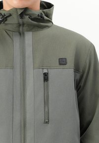 Grøn zip-up jakke med hætte, der har kontrasterende grå paneler, en brystlomme med lynlås og et logo-patch på venstre side.