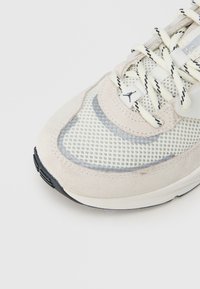 Αθλητικό παπούτσι με επάνω μέρος από μπεζ σουέντ και δίχτυ, λευκές και γκρίζες λεπτομέρειες, με υφασμένο καουτσούκ σόλα και μαύρες λεπτομέρε ιες στη σόλα.