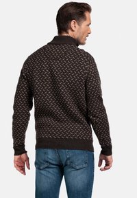 Man in een donkerbruin gebreid patroontrui en blauwe jeans, met zijn rug naar de camera, tegen een effen witte achtergrond.