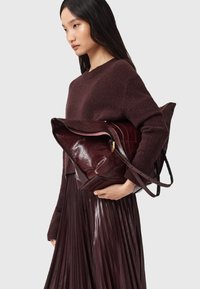 Borsa in pelle borgogna con una forma strutturata, finitura lucida e chiusura a ribalta. Presenta una lunga tracolla e delicati dettagli in oro.