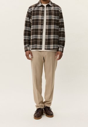 Man wearing brown and white plaid button-up shirt over white tee, beige trousers, and dark brown leather shoes, standing against plain background.