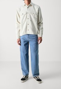 Crème kleurige flanellen shirt met een knoopsluiting, twee borstzakken en gouden knopen, gecombineerd met lichtblauwe jeans en zwarte sneakers.