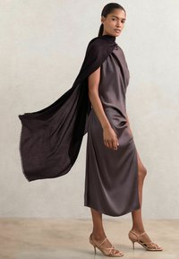 Rochie din satin de lungime până la genunchi, de culoare taupe închis, cu gât drapat și fante laterale, asortată cu o capa neagră fluidă și sandale bej cu curele. Textură netedă.