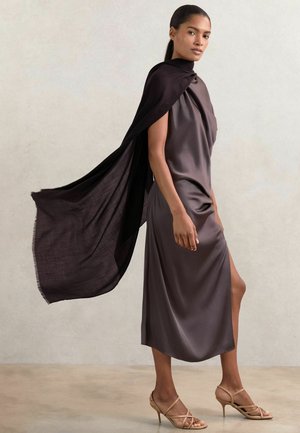 Knee-length, temno taupe saten obleka z drapirano ol neckline in stranskim razporkom, v kombinaciji s flowing črnim plaščem in bež sandali na vezi. Gladka tekstura.