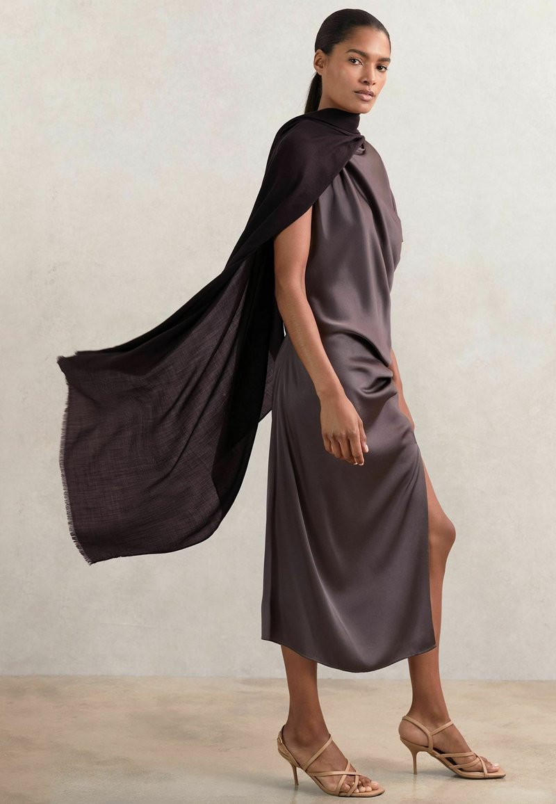 Rochie din satin de lungime până la genunchi, de culoare taupe închis, cu gât drapat și fante laterale, asortată cu o capa neagră fluidă și sandale bej cu curele. Textură netedă.