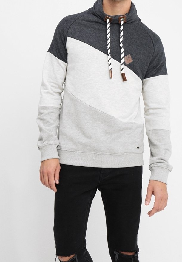 Mężczyzna w bluzie z kapturem w kolorowych blokach: ciemnoszary, jasnoszary i kremowy, w połączeniu z czarnymi podartymi dżinsami.