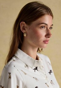 Mladá žena s svetlohnedými vlasmi, ktorá má na sebe zlaté náušnice v tvare kvapky a bielu košeľu s malými potlačami psíkov, hľadí doprava s neutrálnym výrazom.