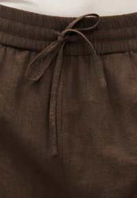 Pantalon en tissu marron avec une ceinture élastique et un cordon de serrage noué au centre devant.