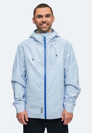 Uomo sorridente che indossa una giacca con cappuccio azzurro chiaro con cerniera blu e pantaloni neri, in piedi davanti a uno sfondo chiaro uniforme.