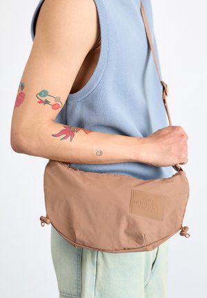 Persoon die een blauwe mouwloze top en lichte denim broek draagt, met kleurrijke tatoeages op de arm, en een beige The North Face crossbodytas vasthoudt.