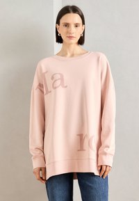 Svetlo ružový oversized sveter s okrúhlym výstrihom, s jemným grafickým potlačou, dlhými rukávmi a voľným strihom. V kombinácii s modrými džínsami.