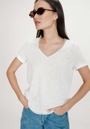 Femme aux longs cheveux bruns portant des lunettes de soleil rondes marron, des boucles d'oreilles argentées en forme d'anneau, un T-shirt blanc à col en V avec une bordure dorée, et un jean bleu.