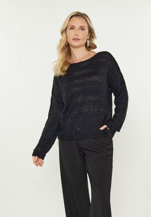 Pulover negru din tricot, cu o croială lejeră, decolteu larg și un model texturat. Asortat cu pantaloni negri, scoțând în evidență umerii căzuți și manșetele ribelate.