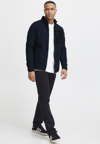 Marineblå fleecejakke med høy krage, glidelås lomme på brystet og svarte detaljer, kombinert med svarte bukser og stripete joggesko.
