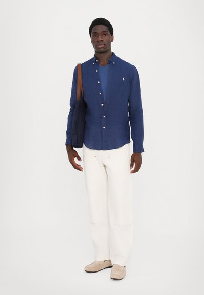 Marineblå lin skjorte med knapper over en blå V-hals, kombinert med vide hvite bukser og beige slip-on sko, holder en mørk totebag.