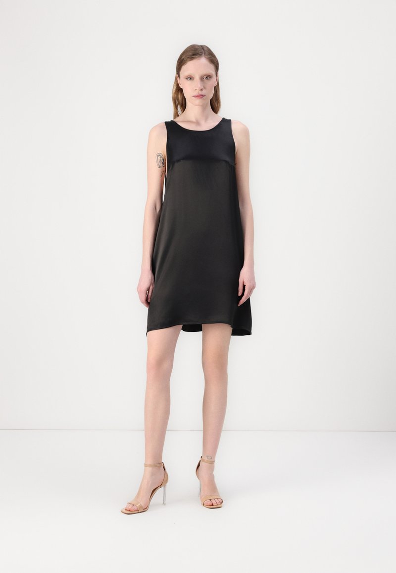 Rochie neagră din mătase, fără mâneci, cu un croi drept, având o textură netedă și un design minimalist. Purtată cu sandale cu toc bej.