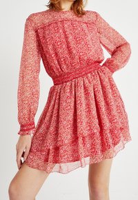 Robe florale rouge en tissu transparent. Elle présente des manches longues, une taille smockée et une jupe fluide avec un ourlet légèrement superposé.