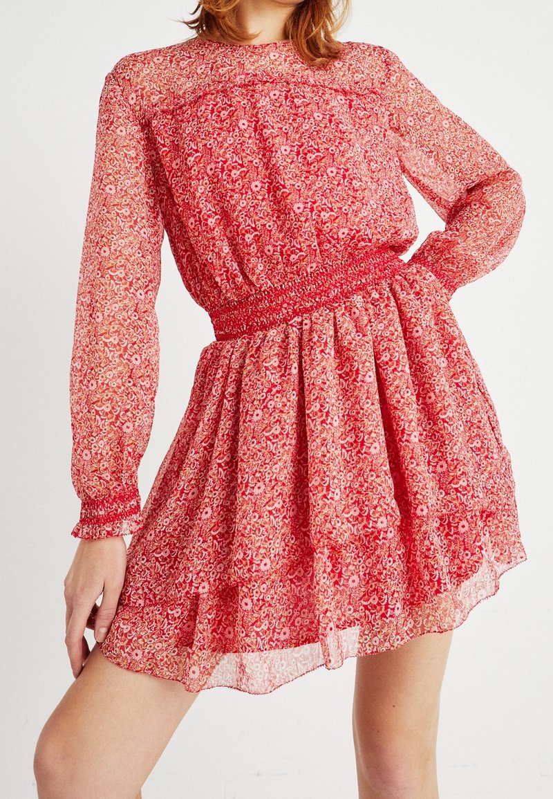Robe florale rouge en tissu transparent. Elle présente des manches longues, une taille smockée et une jupe fluide avec un ourlet légèrement superposé.