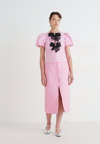 Kvinne iført en pastellrosa topp med pufferm og svarte sløyfer, sammen med et matchende rosa midikjole med frontknapper og slisse, kombinert med sølvfargede sko.