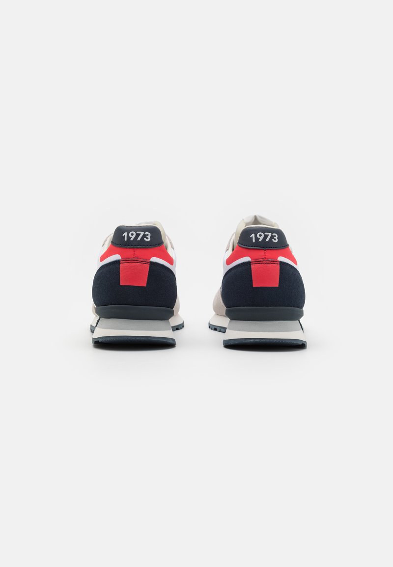 Zapatillas con parte superior beige, acentos en azul marino y detalles en rojo. Presenta un logo con "1973" en el talón y una suela de goma texturizada.